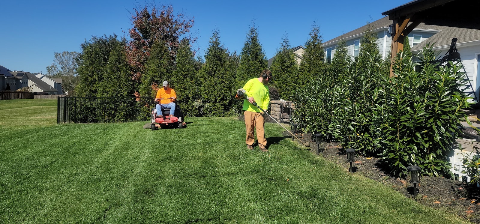 Crew member walking behind a commercial mower across a residential front yard.